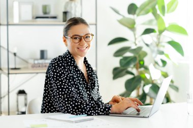 Mutlu beyaz iş kadını gözlük takıyor, parlak modern bir ofiste bilgisayarında yazı yazıyor, bir proje üzerinde çalışıyor, verileri analiz ediyor, notlar alıyor, kameraya gülümsüyor.