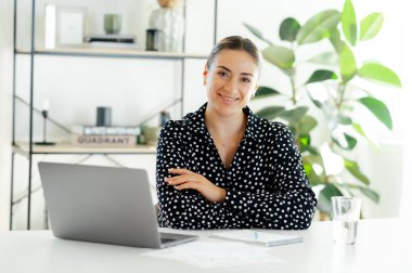 Kendine güveni tam bir iş kadını dizüstü bilgisayarla bir masada oturuyor, kameraya bakıyor ve gülümsüyor, modern ofis alanında başarılı ve motive olmuş hissediyor.
