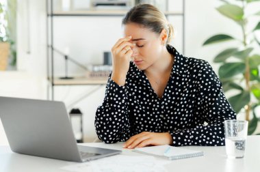 Üzgün, stresli bir iş kadını, ofis sekreteri, gözleri kapalı masada oturuyor, modern bir ofiste şakaklarını ovuyor, dinlenmeye ve dinlenmeye ihtiyacı var. Çok çalışan dişinin migreni var.