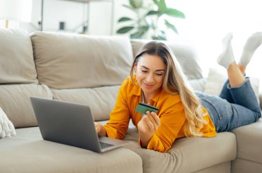 Pozitif beyaz kadın oturma odasındaki kanepede uzanıyor, bilgisayarını kullanırken kredi kartına bakıyor, online alışverişinden ve ödemelerinden memnun ve mutlu görünüyor.