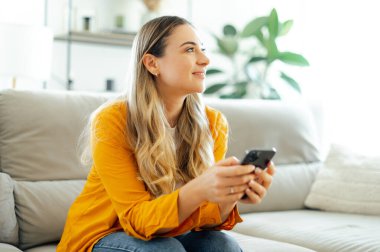 Güzel bir kadın kanepede oturuyor, elinde akıllı bir telefonla, rahat bir ev ortamında cep telefonunu kullanırken mutlu görünüyor, sohbet ediyor veya sosyal medyada geziniyor, başka yere bakıyor ve gülümsüyor.
