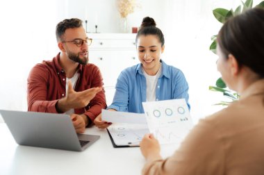 Ofiste takım çalışması konsepti. Bir erkek ve bir kadın, üçüncü bir meslektaşla birlikte mali grafikleri analiz ediyor, belgeler ve dizüstü bilgisayardaki verileri inceliyor, sonuçları tartışıyorlar.