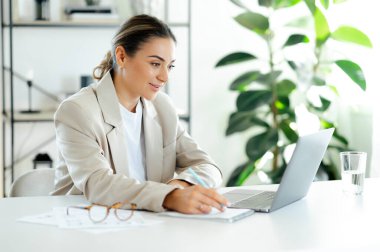 Kafkasyalı bir iş kadını çalışma masasında oturmuş, finansal belgelerle çevrili, gülümseyen modern bir ofiste dizüstü bilgisayarla çalışırken notlar yazıyor.