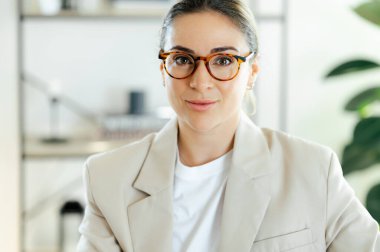 Gözlüklü, ciddi bir ifadeyle büyüleyici başarılı beyaz iş kadınının yakın plan fotoğrafı, doğrudan kameraya bakıyor, modern ofiste güven ve profesyonelliği gösteriyor.