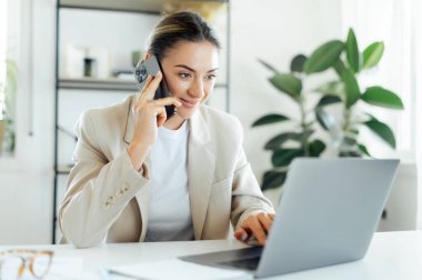 Kendinden emin beyaz bir iş kadını, bej ceketli bir şirket sekreteri iş arkadaşı ya da müşterisiyle telefonda konuşurken modern bir ofiste dizüstü bilgisayarda çalışırken gülümsüyor.