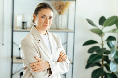 Bej ceketli başarılı beyaz bir iş kadınının portresi modern ofiste kolları çapraz duruyor, kendinden emin görünüyor, liderlik, başarı ve profesyonelliği temsil ediyor.