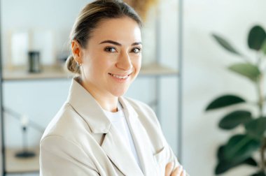 Profesyonel beyaz iş kadınının yakın plan fotoğrafı, kendinden emin ve dostane bir ifadeyle gülümsüyor, başarıyı, liderliği ve kariyer gelişimini temsil ediyor, ofiste duruyor, gülümsüyor.