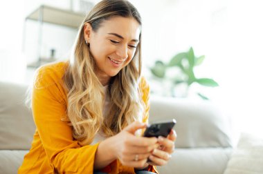 Cihazları kullanarak. Günlük giysiler içinde neşeli beyaz bir kadın oturma odasındaki bir kanepede rahat bir şekilde otururken akıllı telefonunu kullanıyor. Gülümsüyor, ekranlara bakıyor, çevrimiçi bir sohbetin tadını çıkarıyor.