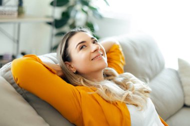 Sarı tişörtlü, mutlu, sakin, beyaz kadın rahat bir koltukta uzanmış, mutlu bir gülümsemeyle yukarıya bakıyor, rahat evinde huzurlu ve mutlu bir hava sergiliyor.