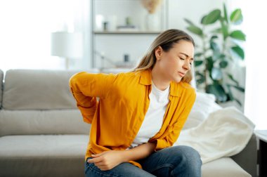 Sırt ağrısı kavramı. Evdeki bir kanepede gözleri kapalı oturan, sırtı ağrılı bir ifadeyle tutan, sırt ağrısı ya da omurga rahatsızlığından muzdarip beyaz kadın.