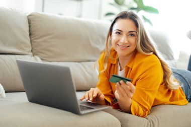 İnternetten alışveriş, elektronik bankacılık. Sarı gömlekli bir kadın evdeki kanepede uzanmış, elinde bir kredi kartıyla dizüstü bilgisayarını karıştırıyor, rahat bir evde internetten alışveriş yapıyor.