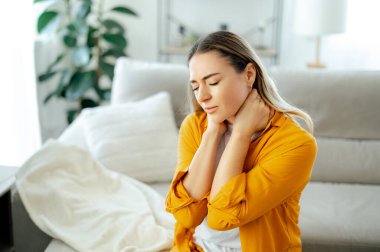 Üzgün, beyaz bir kadın, evinde, gözleri kapalı bir kanepede oturuyor, iki eliyle boynunu tutuyor ve rahatsızlığını dile getiriyor, boyun ağrısı ya da kas gerilimi yaşıyor, dinlenmeye ve masaja ihtiyacı var.