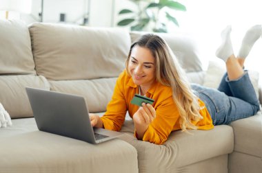 Neşeli beyaz kadın, günlük giysiler içinde, oturma odasındaki rahat bir kanepede yatıyor, dizüstü bilgisayarına bakarken bir kredi kartı tutuyor, online alışveriş yapıyor, faturaları ödüyor, sipariş veriyor, gülümsüyor.