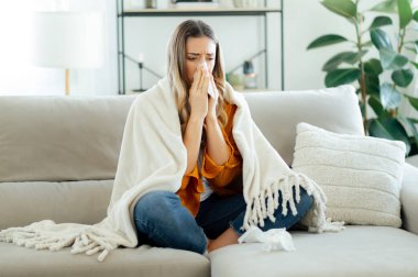 Sarı gömlekli sağlıksız beyaz kadın oturma odasında yumuşak beyaz bir battaniyeye sarılmış, burnunu mendile sümkürmüş, iyi görünmüyor, nezle ya da grip belirtileri gösteriyor.