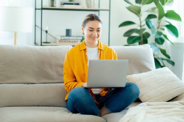 Sarı tişörtlü, kot pantolonlu, bej koltukta bağdaş kurmuş, dizüstü bilgisayarda çalışan beyaz bir kadın. Gülümseyerek ekrana bakıyor. Oturma odasında online bir iş yapıyor.