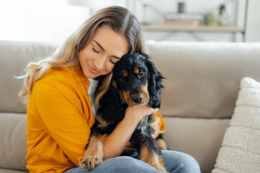 Sarı gömlekli, uzun sarı saçlı, mutlu, beyaz bir kadın, rahat bir oturma odasında kanepede oturur, siyah ve kahverengi köpeğine sevgi dolu bir ifadeyle sarılır.