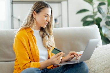 Sarı tişörtlü, uzun beyaz bir kadın oturma odasında bir kanepeye oturur ve dizüstü bilgisayar kullanır. Elinde bir kredi kartı tutar ve online alışverişe odaklanır.