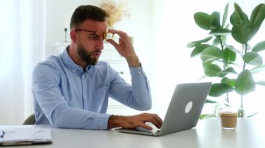 Mavi gömlekli düşünceli bir adam laptopuyla bir masada oturuyor, etrafı çizelgeler ve bir fincan kahveyle çevrili, çalışmalarını yansıtıyor, sonuçlar ve raporlar için endişeleniyor.