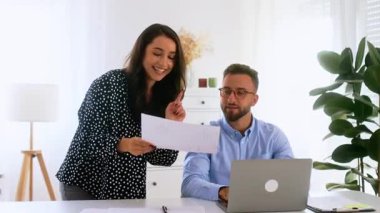 Olumlu iş kadını, modern ofisteki bir erkek meslektaşına iş belgesi gösteriyor. İkisi de gülümsüyor, dizüstü bilgisayarı ve çizelgeleri masada. Fikirleri paylaşıyor, iş planı geliştiriyor ve strateji geliştiriyor. 