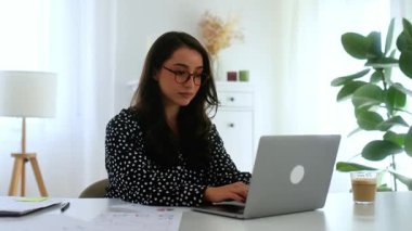 Gözlüklü güzel esmer iş kadını, gömlek giymiş, şirket yöneticisi ya da tasarımcısı, masa başında oturmuş, parlak bir ofiste evraklarla çevrili, kameraya gülümsüyor.