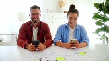 Gadgets konsepti. İki meslektaş, erkek ve kadın, ofiste bir masada oturuyorlar, akıllı telefonlarını kullanıyorlar, müşterileri ve arkadaşlarıyla mesajlaşıyorlar, haberleri okuyorlar, bilgi arıyorlar, kameraya gülümsüyorlar.
