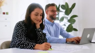 Mutlu, çekici, genç bir Arap ya da beyaz bir kadın masadaki yapışkan bir nota yazı yazıp kameraya gülümserken bir adam arka planda dizüstü bilgisayara yazarak takım çalışması ve yaratıcılık sergiliyor.