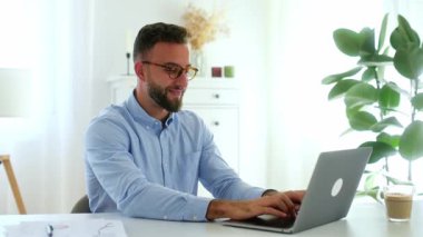 Başarı golü. Mavi gömlekli neşeli başarılı iş adamı, dizüstü bilgisayarına bakarken yumruklarını kaldırıyor, parlak, modern bir ofiste başarısını kutluyor, mutlu bir şekilde gülümsüyor.