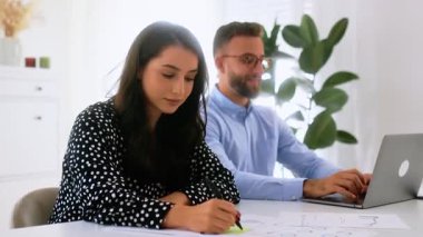 Neşeli, genç, başarılı esmer iş kadını, parlak, modern bir ofiste masa başında çalışıyor, kameraya gülümsüyor, erkek arkadaşı ise arka planda dizüstü bilgisayara yazılar yazıyor.