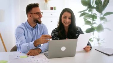 Takım çalışması konsepti. Modern bir ofiste belgelerle çalışan iki dostane iş ortağı. İkisi de gülümsüyor, dizüstü bilgisayarları ve çizelgeleri var. Fikirlerini paylaşıyorlar, iş planı geliştiriyorlar.