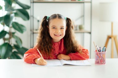 Mavi gözlü mutlu bir kız 6-7 yaş arası, kırmızı bir bluz giymiş, kıvırcık saçlı, elinde kalemle evde oturuyor ve ödevini yaparken kameraya gülümsüyor.