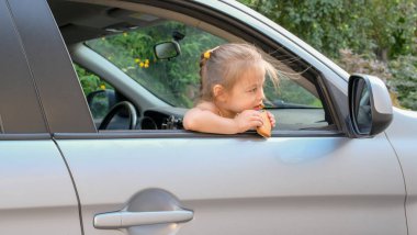 Arabasında oturan, pencereden dışarı bakan ve yemek yiyen sevimli küçük bir kız. Arabayla seyahat eden küçük bir çocuk. Çocuk güvenliği yolda. Aile gezisi ve yaz tatilleri. Yüksek kalite