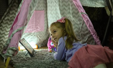 Küçük kız oyun evinde battaniyeye uzanmış, tabletteki çizgi filmi ilgiyle izliyor. Yüksek kalite fotoğraf