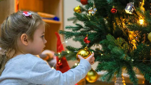 一个穿着裙子的迷人的小女孩在圣诞树上挂着一个闪闪发光的球 一个孩子正在漂亮的客厅里装饰圣诞树 家庭节日快乐的概念 免版税图库照片