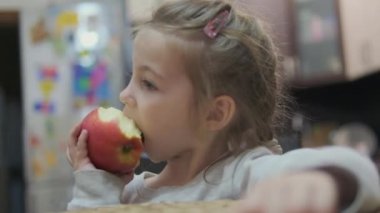 Küçük kız mutfakta masada oturur ve büyük sulu bir kırmızı elma yer. Yüksek kaliteli FullHD görüntüler
