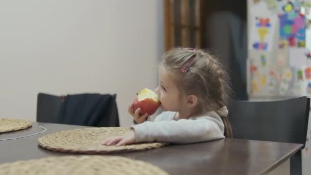 小さな女の子は台所でテーブルに座り 大きなジューシーな赤いリンゴを食べる 高品質のフルHd映像 — ストック動画