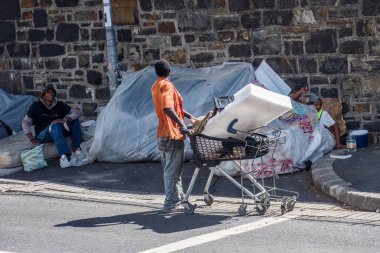 Cape Town, Güney Afrika - 2 Nisan 2024: Turuncu tişörtlü evsiz bir adam asfalt yüzey boyunca içi çeşitli eşyalarla dolu bir alışveriş arabasını itiyor