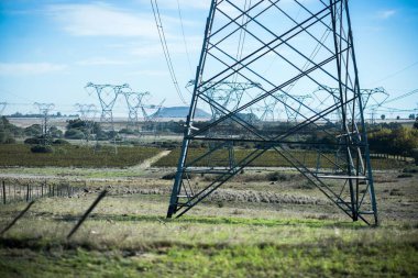 Yüksek voltajlı elektrik hatlarından oluşan bir ağ, elektrik kablolarını destekleyen uzun metal kuleleriyle kırsal bir araziye yayılır. Ön plandaki geniş açık alanlar, geniş mavi alanların altında, altyapıların endüstriyel varlığıyla tezat oluşturuyor. 