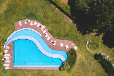 Çimen ve ağaçlarla çevrili kavisli bir yüzme havuzunun havadan görünüşü. Havuz güvertesinde birkaç beyaz şezlong var.