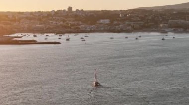 Bir yelkenli gün batımında Cascais limanının sakin sularında yol alır. Güneş şehrin arkasında batıyor, suyun üzerine altın bir parıltı saçıyor.