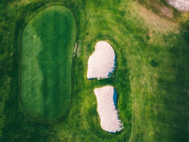 Bir golf sahasının yeşili yukarıdan belirgin bir şekilde gösterilir ve pürüzsüz yüzeyini gösterir. Yeşilliğin yanında, bir kum sığınağı görülebilir durumda. Çevredeki çimenlere açıkça çizilmiş.. .