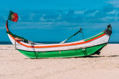 Parlak boyalı bir moliceiro teknesi kumlu sahilde Portekiz bayrağı sallıyor. Huzurlu okyanus ve canlı gökyüzü sakin bir kıyı atmosferi yaratır.