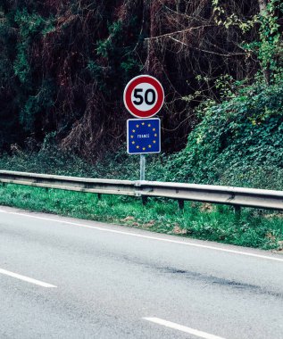 Fransa 'da kırsal bir yolda gündüz vakti yeşillik ve ağaçlarla çevrili saatte 50 km hız sınırı işareti var.