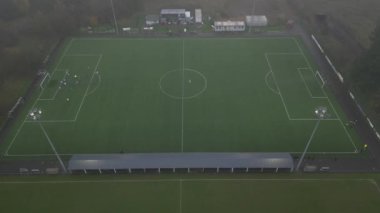 Gri ve yağmurlu bir günde yapılan futbol antrenmanına genel bir bakış. Oyuncular aktif olarak antrenman yaparken, koçlar saha kenarından izliyor.
