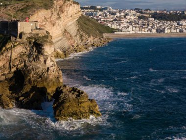 Nazare, Portekiz 'deki deniz fenerinden gelen dev dalgalara hayran olan insanların insansız hava aracı görüntüsü güneşli bir günde en sağdaki Nazare kasabasıyla birlikte Kuzey Sahili' ne bakıyor.