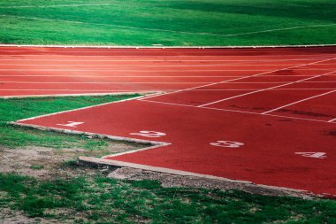 Boş bir atletizm pistinde, taze yeşil çimlerle çevrili sayılı şeritler ve canlı kırmızı yüzey yer alıyor, sporcuları sabah antrenmanlarına veya yarışmalarına davet ediyor, adanmışlık ve sportmenlik sergiliyor.