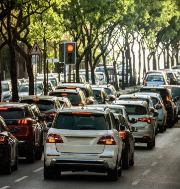 Güneş arka planda batarken, arabalar ağaçlarla kaplı bir cadde boyunca yoğun trafikte dizilmiş ve yaprakların arasından altın ışık saçıyor. Trafik ışığı kırmızı, durdurulan araçları gösteriyor.