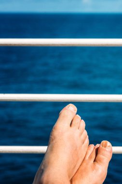 Güzel mavi bir okyanusa bakan beyaz bir korkulukta çıplak ayaklar dinleniyor. Güneş ışıl ışıl parlıyor ve kıyı manzarasının tadını çıkarmak için rahatlatıcı bir atmosfer yaratıyor.