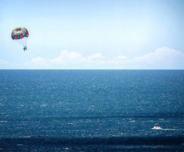 İnsanlar açık bir günde kumlu bir sahil kenarında, aşağıda renkli paraşüt ve tekne olan deniz paraşütlerini severler.