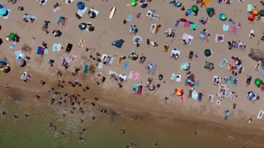 Kıyı kenarında bir yaz öğleden sonrası boyunca kalabalık bir plajı renkli şemsiyeler ve güneş banyosu eşliğinde tepeden aşağıya bakan hava aracı.