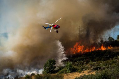 Bir helikopter dağlık bir bölgedeki orman yangınının üzerinde süzülerek alevlerle savaşmak için su bırakıyor. Yoğun duman mavi gökyüzüne yükseliyor. Bu da Portekiz 'de ciddi bir yangın tepkisine işaret ediyor.